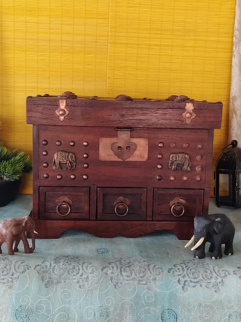 Op de afbeelding: Een donkerbruine houten kist met drie laden en decoratieve olifantenaccenten. De kist heeft een scharnierend deksel met een metalen sluiting en koperen accenten. De kist staat op een blauw-groen gepatroneerd oppervlak.