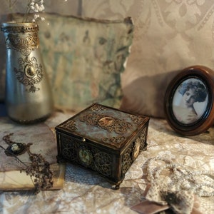 May include: A square, ornate, wooden box with a teal lid and gold accents. The box is decorated with intricate metalwork and has a vintage aesthetic. It is sitting on a table with a vase of white flowers and a framed picture.