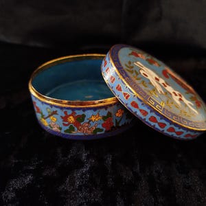 May include: A round, open trinket box with a blue interior and lid. The lid features a white rabbit design with red and gold accents. The box has a gold rim and is set against a dark background.
