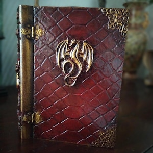 May include: A red leather-bound journal with a gold dragon design on the front cover. The journal has gold accents and a decorative clasp.