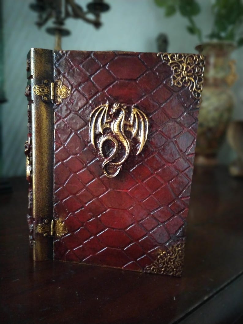 May include: A red leather-bound journal with a gold dragon design on the front cover. The journal has gold accents and a decorative clasp.