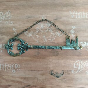 May include: A decorative key-shaped wall hook with a chain for hanging. The key is made of metal and has a green patina finish. The key has a decorative design on the top and a small hook on the bottom.