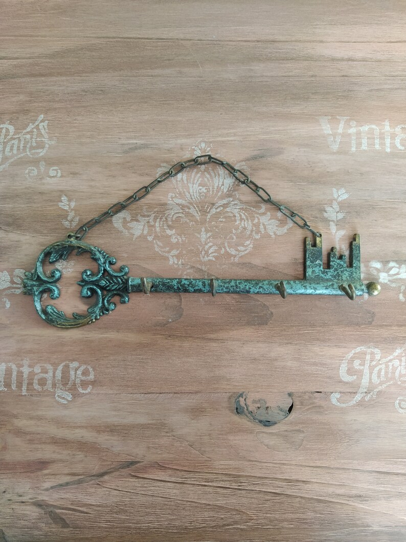 May include: A decorative key-shaped wall hook with a chain for hanging. The key is made of metal and has a green patina finish. The key has a decorative design on the top and a small hook on the bottom.
