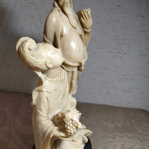 May include: A white ceramic sculpture of a man and woman in traditional Chinese clothing. The man is holding a cup and the woman is holding a bunch of grapes. The sculpture is on a black base.