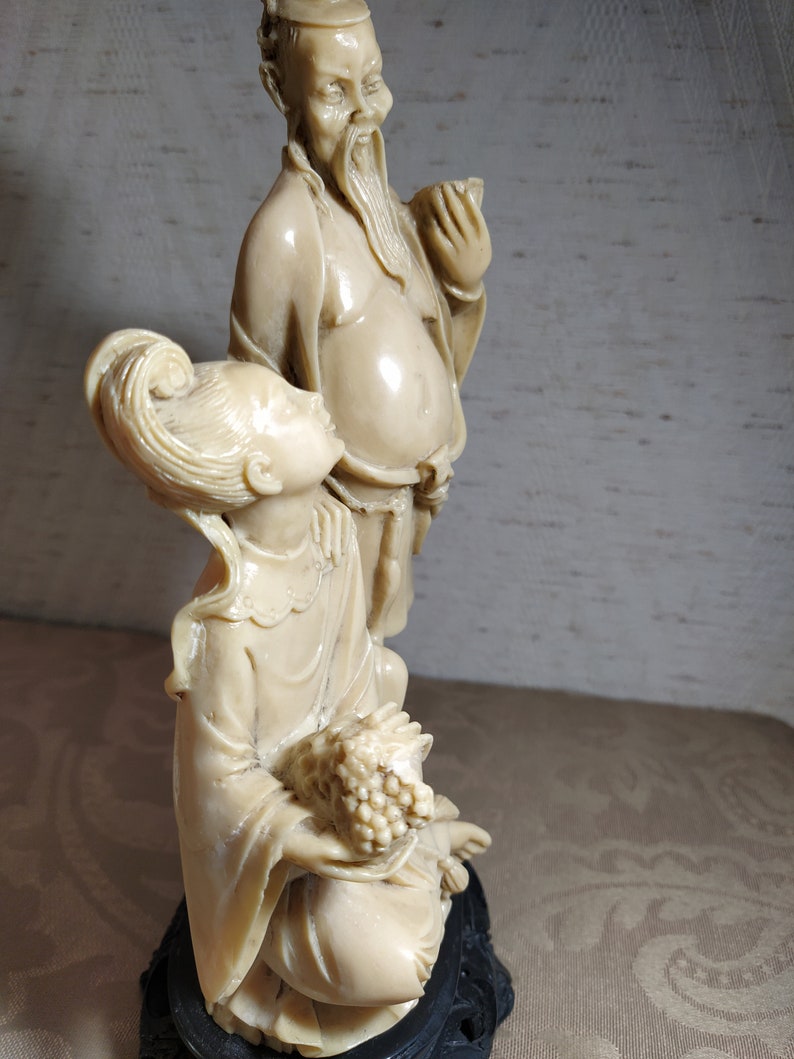 May include: A white ceramic sculpture of a man and woman in traditional Chinese clothing. The man is holding a cup and the woman is holding a bunch of grapes. The sculpture is on a black base.