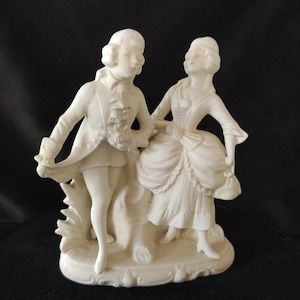May include: A white porcelain figurine of a couple in a romantic pose. The man is wearing a jacket and breeches, and the woman is wearing a dress with a large skirt. The figurine is set on a white base with a decorative design.
