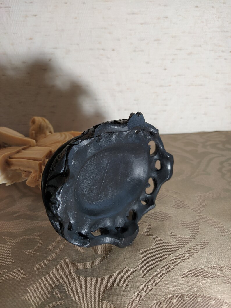 May include: A black carved wooden base with intricate details. The base has a circular shape with a raised edge and a small hole in the center.