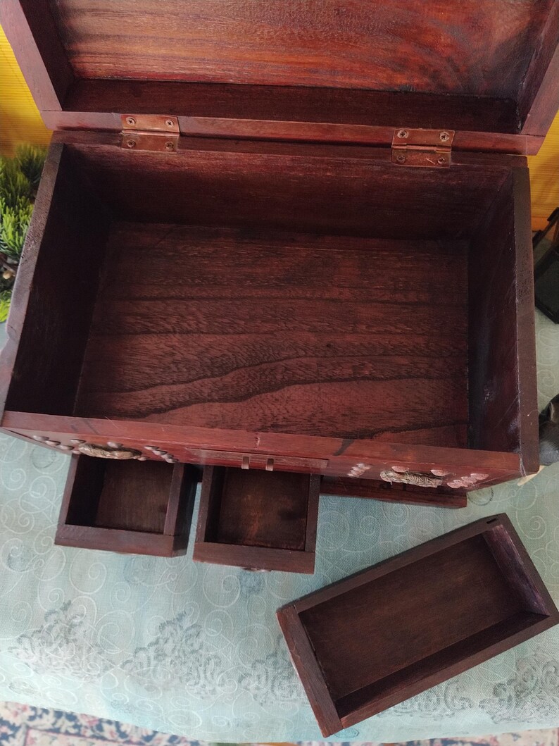 Op de afbeelding: Een houten doos met een scharnierend deksel en drie laden. De doos is gemaakt van donker hout met een natuurlijke afwerking. De laden zijn allemaal open en leeg.