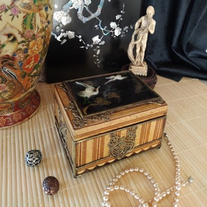 May include: A wooden box with a black and gold design. The top of the box features a painted scene of two white birds flying over a lake. The box is decorated with intricate gold trim and has a striped pattern on the sides.