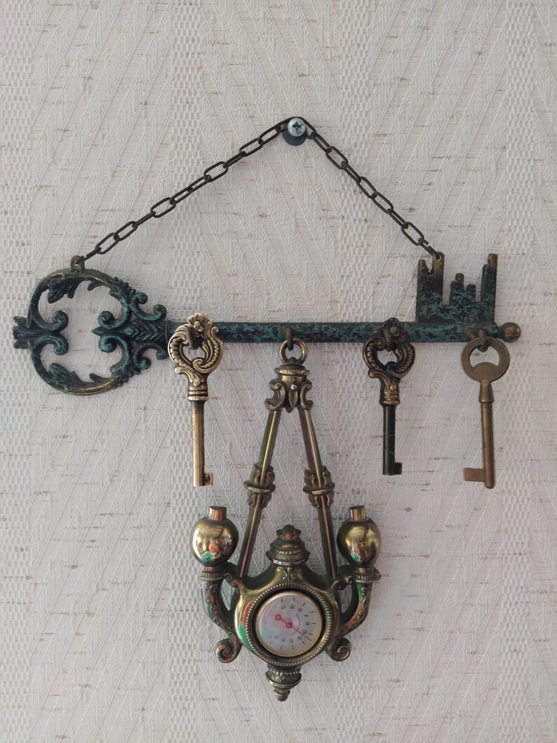 May include: A decorative key-shaped wall hanger with a chain, three keys, and a hanging thermometer with a brass finish.