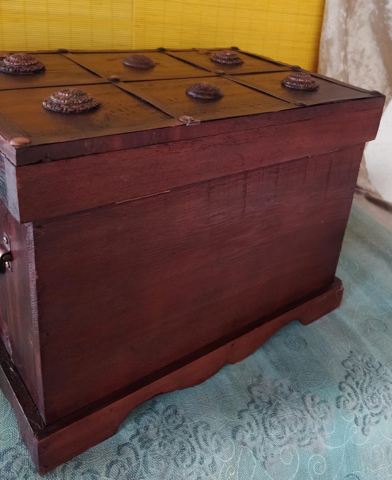 Op de afbeelding: Een donkerbruine houten kist met een scharnierend deksel. Het deksel heeft een decoratief ontwerp met metalen accenten en een textuur.