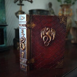 Puede incluir: Un pequeño libro encuadernado en cuero rojo con detalles dorados y un diseño de dragón en la portada. El libro está cerrado y de pie sobre una superficie de madera.