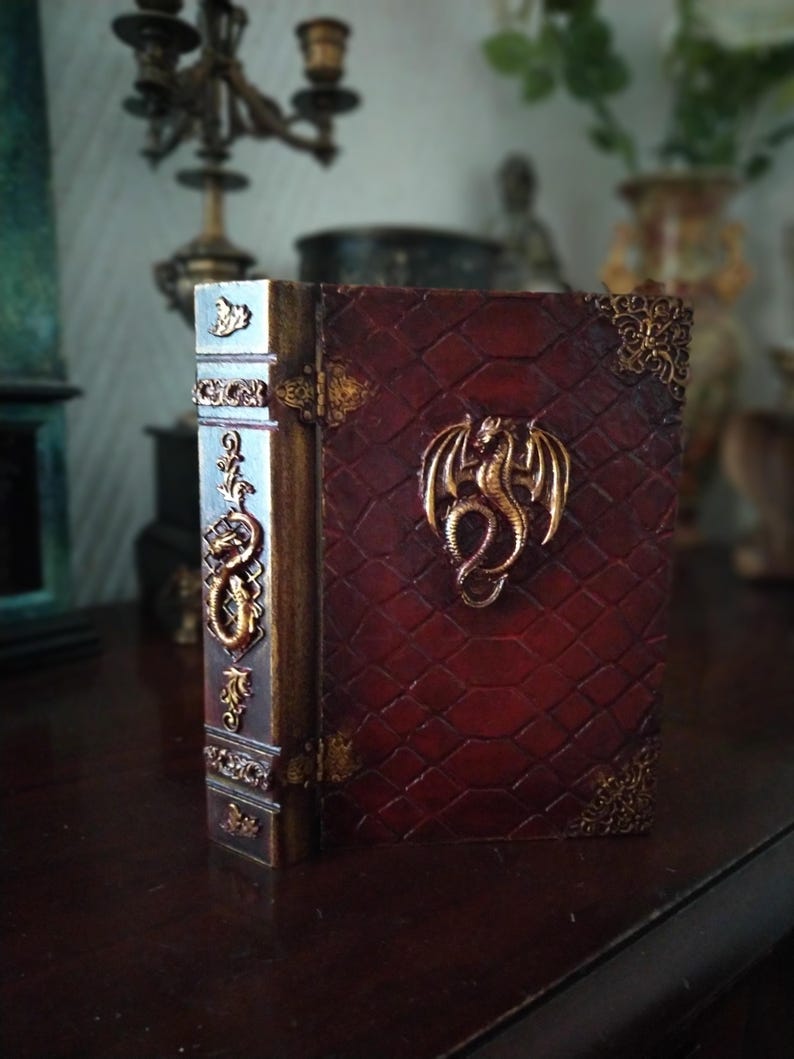 May include: A small, red leather-bound book with gold accents and a dragon design on the front cover. The book is closed and standing upright on a wooden surface.