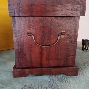 Op de afbeelding: Een donkerbruine houten kist met een scharnierend deksel en een metalen handvat. De kist heeft een decoratieve basis met een gegolfde rand.