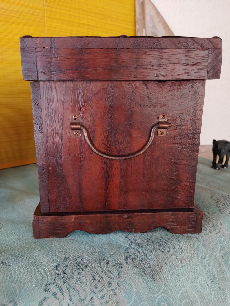 Op de afbeelding: Een donkerbruine houten kist met een scharnierend deksel en een metalen handvat. De kist heeft een decoratieve basis met een gegolfde rand.