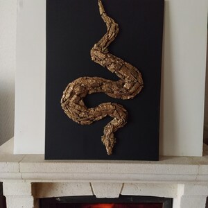 Könnte beinhalten: Eine goldfarbene Schlangenskulptur aus Baumrinde, montiert auf einer schwarzen Leinwand. Die Skulptur ist in einer gewundenen Position, mit Kopf und Schwanz sichtbar.