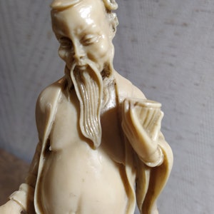 May include: A white ceramic figurine of a man with a long beard and a hat. He is wearing a long robe and holding a scroll in his hand.