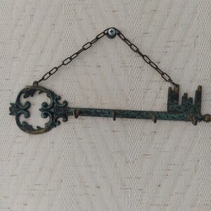 May include: A decorative key-shaped wall hook with a chain and a screw for hanging. The hook is made of metal and has a distressed green finish. It features a large ornate keyhole and five hooks on the other end.
