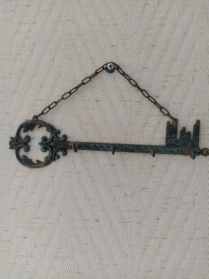 May include: A decorative key-shaped wall hook with a chain and a screw for hanging. The hook is made of metal and has a distressed green finish. It features a large ornate keyhole and five hooks on the other end.
