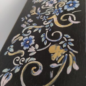 以下が含まれることがあります： 黒色の木製の表面に手描きの花の模様が施されています。模様には青、白、金色の花と蔓が描かれています。