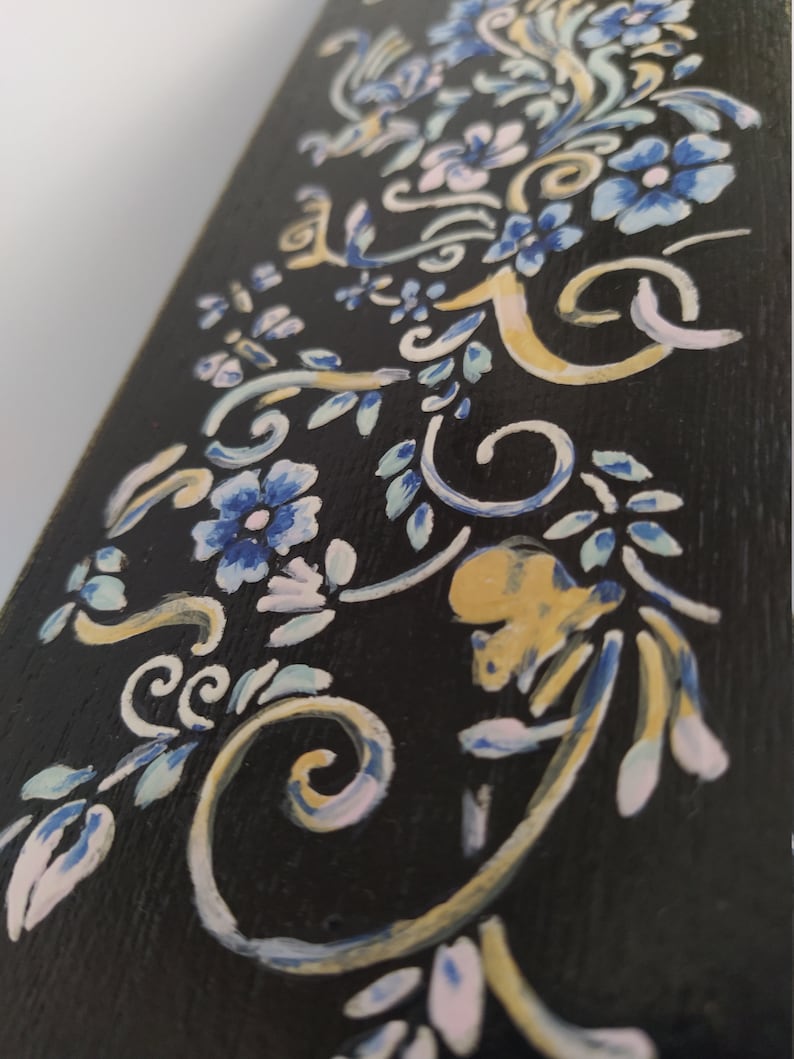 以下が含まれることがあります： 黒色の木製の表面に手描きの花の模様が施されています。模様には青、白、金色の花と蔓が描かれています。