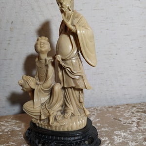 May include: A cream-colored ceramic sculpture of two figures in traditional Chinese clothing. The figures are standing on a black base with intricate details. The sculpture is a traditional art form.