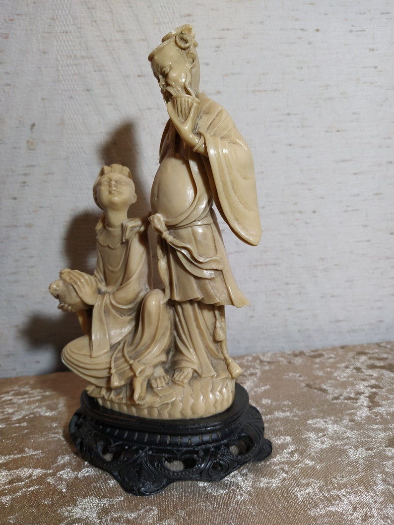 May include: A cream-colored ceramic sculpture of two figures in traditional Chinese clothing. The figures are standing on a black base with intricate details. The sculpture is a traditional art form.