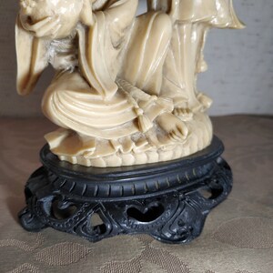 May include: A cream-colored ceramic statue of two figures, one seated and holding a round object, the other standing. The statue is on a black base with intricate carvings.