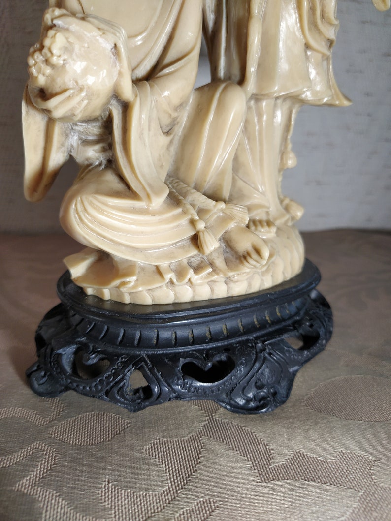 May include: A cream-colored ceramic statue of two figures, one seated and holding a round object, the other standing. The statue is on a black base with intricate carvings.