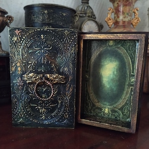 May include: A dark green wooden box with a gold-colored ornate design and a brass handle. The box is open, revealing a green and gold interior with a mirror.
