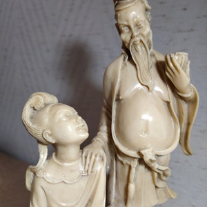 May include: A pair of ivory-colored figurines depicting a man and a woman in traditional Chinese clothing. The man is holding a small object in his hand and the woman is looking up at him.