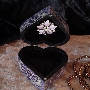 May include: Open heart-shaped jewellery box with a floral brooch inside. The box has a black interior and a purple and white patterned exterior. A beaded necklace lies beside the box. The brooch is pale pink with clear crystal accents.