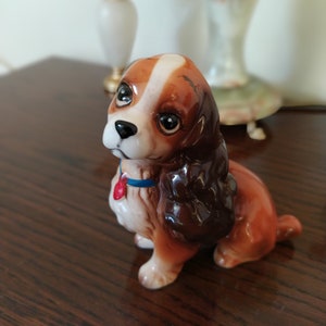 May include: A ceramic figurine of Lady, the cocker spaniel from Disney's Lady and the Tramp. The figurine is brown and white with a blue collar and a red heart charm. The figurine is sitting on a wooden surface.
