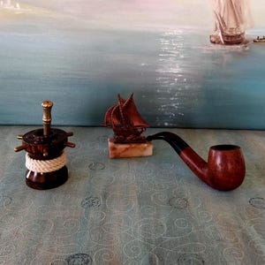 May include: A selection of nautical-themed items. Features a dark brown wooden pipe, a decorative ship sculpture on a marble base, and a dark brown wooden bottle stopper with a gold handle and rope detail.