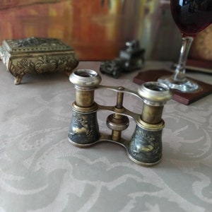 May include: Antique silver and black opera glasses with intricate engravings. The glasses are closed and sitting on a patterned tablecloth.