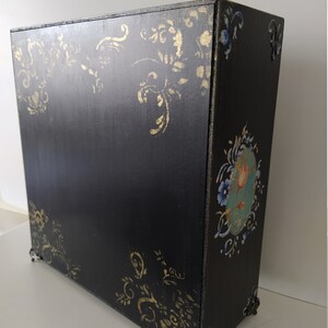 以下が含まれることがあります： 金色と青色の花柄が施された黒い木製の箱。箱には装飾的な金属製の脚と蝶番付きの蓋が付いています。