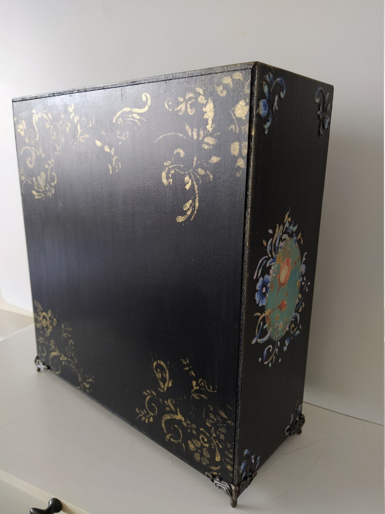 以下が含まれることがあります： 金色と青色の花柄が施された黒い木製の箱。箱には装飾的な金属製の脚と蝶番付きの蓋が付いています。