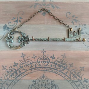 May include: A key-shaped metal key holder with a chain hanging from the top. The key holder has a decorative design and is painted in shades of green and brown. The key holder is mounted on a wooden surface with a decorative pattern.