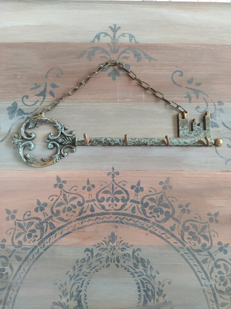 May include: A key-shaped metal key holder with a chain hanging from the top. The key holder has a decorative design and is painted in shades of green and brown. The key holder is mounted on a wooden surface with a decorative pattern.
