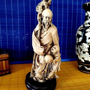 May include: A beige ceramic figurine of a man with a long beard holding a staff and a child. The figurine is standing on a wooden base.