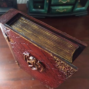 May include: A red book-shaped box with a gold-colored dragon design on the front. The box has a gold-colored spine and a decorative gold-colored border.
