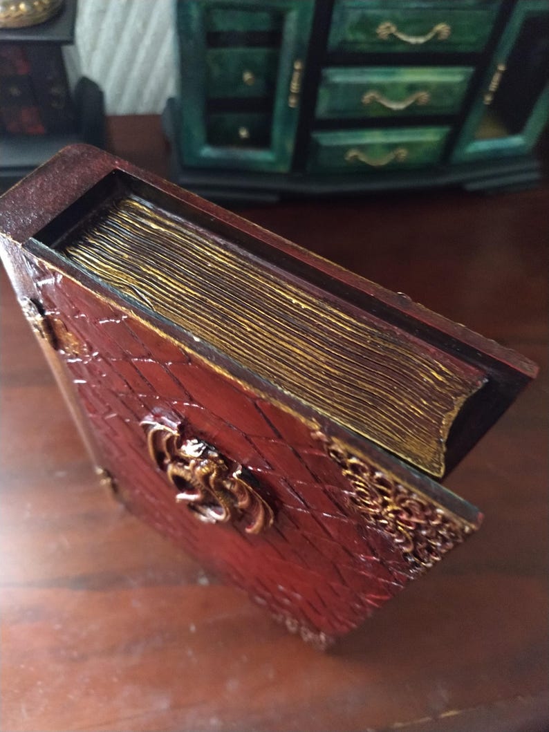 May include: A red book-shaped box with a gold-colored dragon design on the front. The box has a gold-colored spine and a decorative gold-colored border.