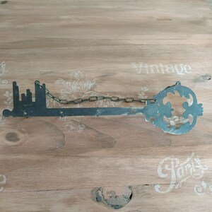 May include: A large, black metal key with a chain attached. The key is shaped like a city skyline and has a decorative flourish at the end. The key is lying on a wooden surface with the words "Vintage" and "Paris" printed on it.