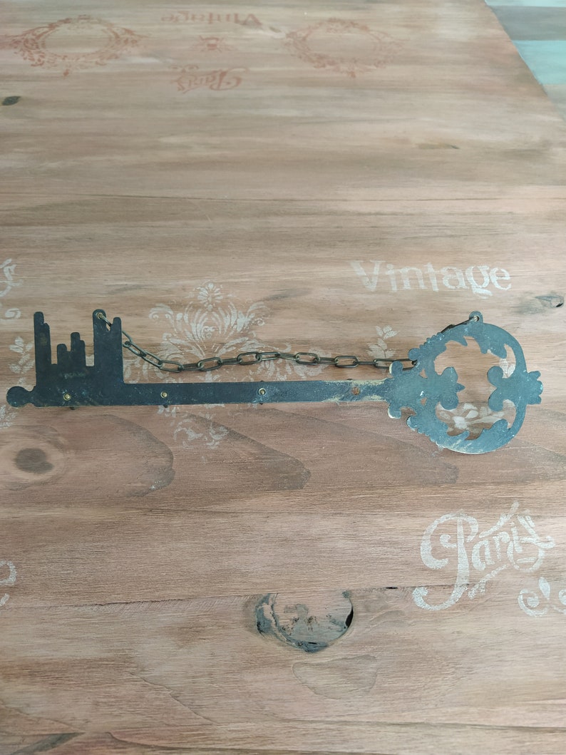 May include: A large, black metal key with a chain attached. The key is shaped like a city skyline and has a decorative flourish at the end. The key is lying on a wooden surface with the words "Vintage" and "Paris" printed on it.