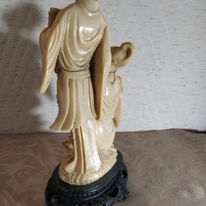 May include: A cream-colored ceramic figurine of two people standing on a black base. The figurine is carved in a traditional style and depicts a man and a woman in flowing robes.
