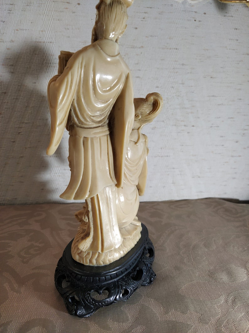 May include: A cream-colored ceramic figurine of two people standing on a black base. The figurine is carved in a traditional style and depicts a man and a woman in flowing robes.