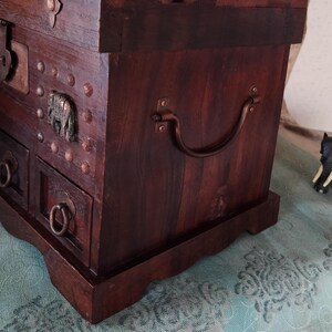 Op de afbeelding: Een donkerbruine houten kist met een metalen handvat en decoratieve metalen accenten. De kist heeft een scharnierend deksel en meerdere laden.