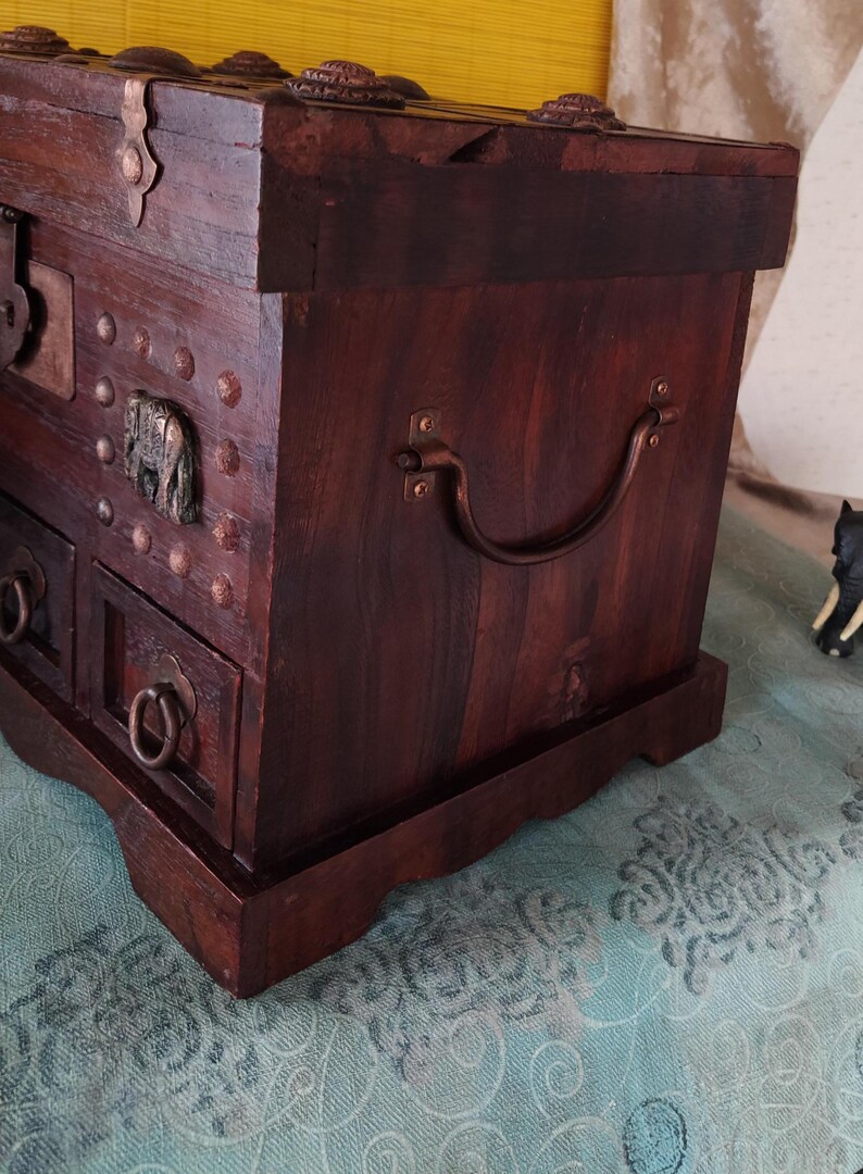 Op de afbeelding: Een donkerbruine houten kist met een metalen handvat en decoratieve metalen accenten. De kist heeft een scharnierend deksel en meerdere laden.