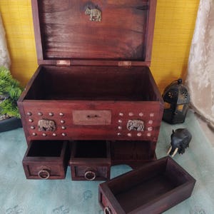 Op de afbeelding: Een donkere houten juwelendoos met drie laden en een opklapbaar deksel. De doos is versierd met metalen olifantenaccenten en studs. De doos is open en de laden zijn uitgetrokken.
