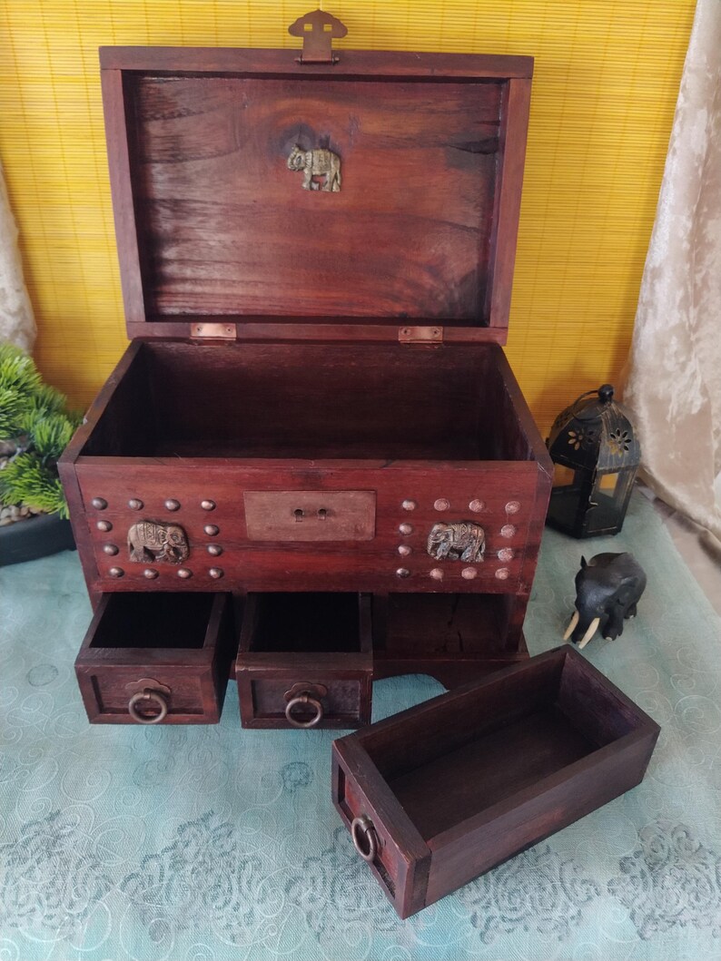 Op de afbeelding: Een donkere houten juwelendoos met drie laden en een opklapbaar deksel. De doos is versierd met metalen olifantenaccenten en studs. De doos is open en de laden zijn uitgetrokken.